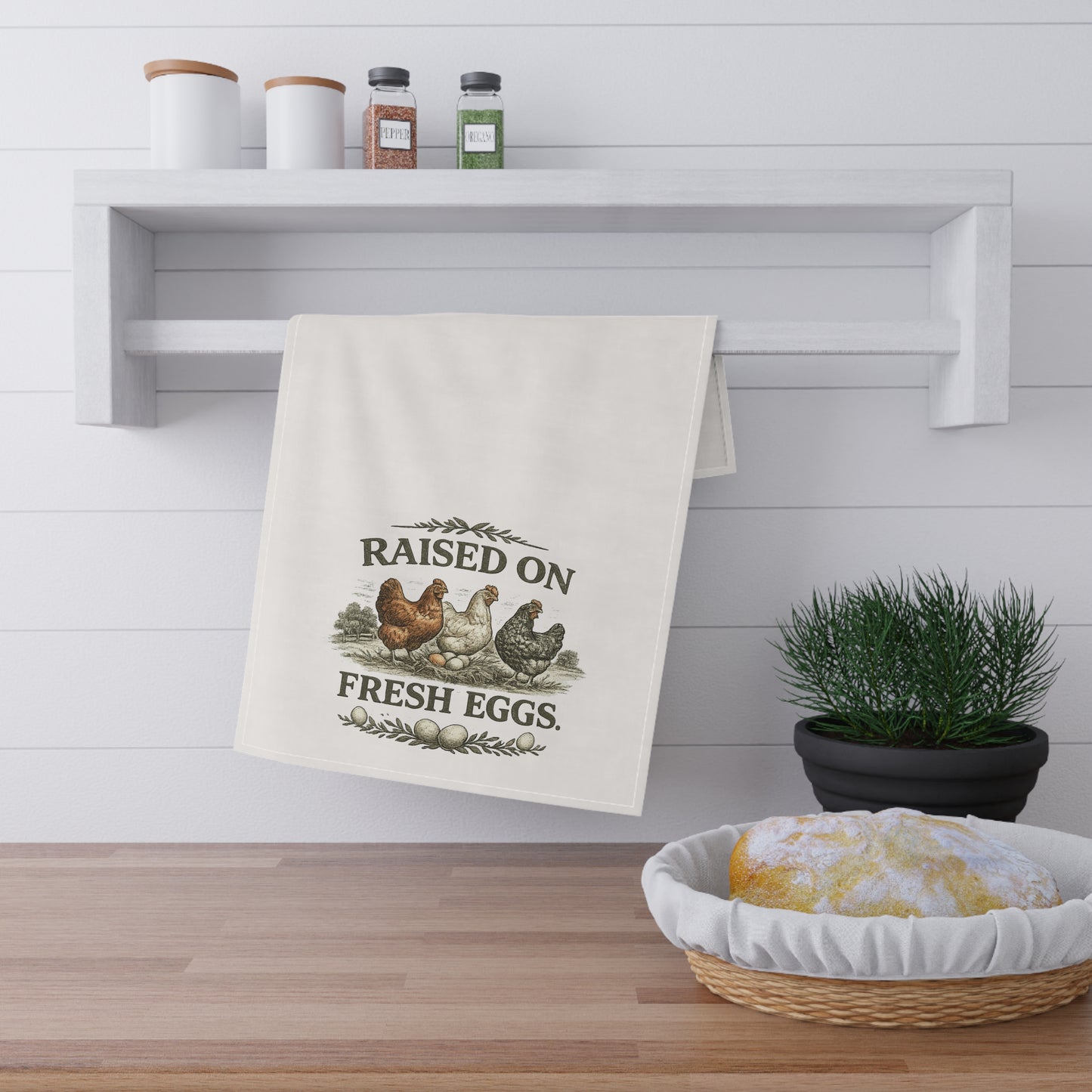 Kitchen towel with 'Raised on Fresh Eggs' text and chicken graphics hanging on a white wall above a wooden counter.
